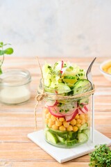 Healthy salad of cucumber, boiled chickpeas, radish, iceberg lettuce and microgreens in a glass jar on a wooden background. Healthy food, diet, vegan concept.