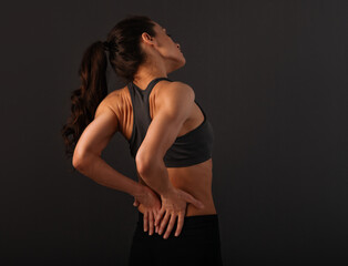 Female sporty muscular model with ponytail doing correction massage holding two hands on the low back on dark shadow grey background with empty copy space.