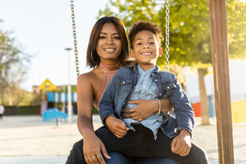 African brothers are in an outdoor park on a swing. The boy with the afro hair is on top of the teenage girl while they look at the camera happily. Concept of brotherly love.