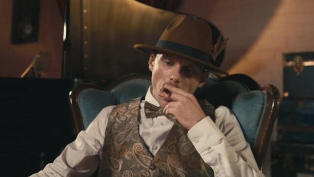 Close up shot of young confident man with mustache, in classy dandy style hat and elegant gentlemen outfit. Guy smoking cigar and starting funk style dance in studio with art deco victorian interior