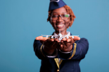 Funny flight attendant in uniform landing model airplane in hands, playing with small custom toy. Airplane crew member with passenger toy, studio shot against blue background.