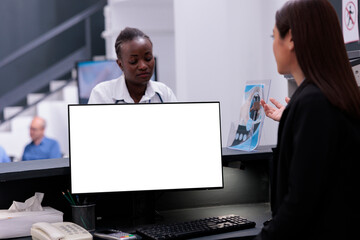 Asian receptionist using white screen on computer while working at hospital reception desk. Worker looking at isolated mockup template while talking with doctor during checkup visit