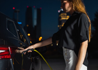 Young modern woman charging an electric EV car in city at night