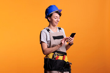 Happy female contractor browsing internet on mobile phone app, reading social media text over background. Cheerful professional builder using smartphone app or network in studio.