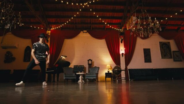 Guy breakdancing street hiphop dance. Dancing man in stylish hat performing floor spinning freestyle dance indoors in victorian style classic hall having fun. Vintage retro lifestyle happiness concept