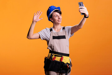 Happy female builder talking on online video call with smartphone over yellow background, using...