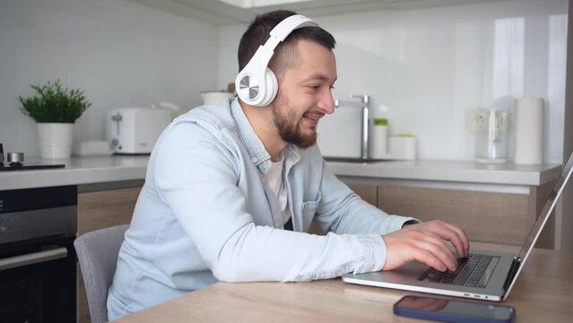 happy smiling caucasian male student wearing headphone sit at kitchen table, watching online video conference learning virtual course teaching using laptop computer in homeoffice.