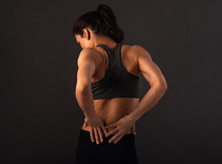 Female sporty muscular model with ponytail doing correction massage holding two hands on the low back on dark shadow grey background with empty copy space. - 599413906