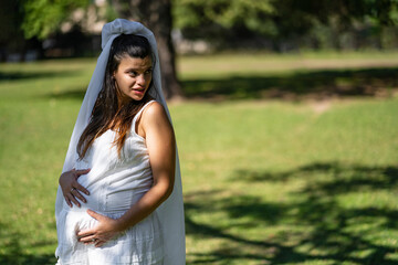 Latin pregnant bride in a white wedding dress in a park. Happy expression