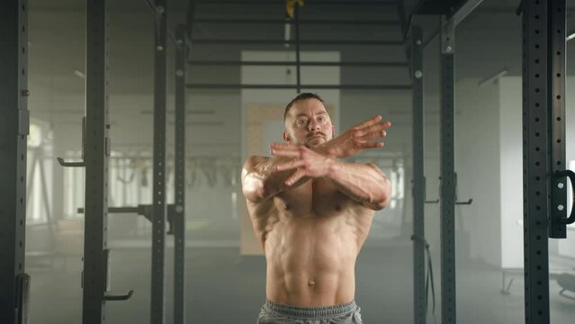 Middle-aged bodybuilder with bare fit torso posing in front of camera standing in power rack. Man keeps hands on waist, raises hands up, shows muscles. High quality 4k footage