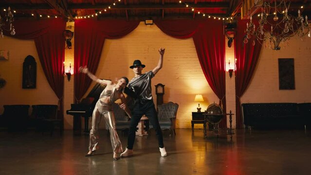 Fashionable girl acrobat performs flips with twine, doing cartwheel supported by dance partner. Slow motion pair of dancers training in retro style interior with chandeliers cinematic RED camera shot