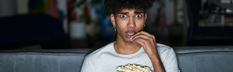 Frightened young guy with eyes wide open wearing pajamas eating popcorn while watching horror...