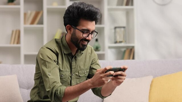 An Indian man is sitting on the sofa in his living room and playing an online game on his mobile phone, raising his hands up in victory celebration.
