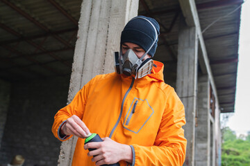 One young guy drawing graffiti on wall with spray can and mask on his face during the day