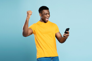 Happy African American man holding  mobile phone sports betting, win money isolated on blue background. Overjoyed gambler playing mobile game celebration success. Happy guy shopping online with sale  - obrazy, fototapety, plakaty