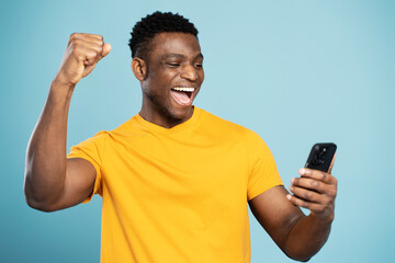 Overjoyed African man using smartphone sports betting, win money isolated on blue background. Emotional Nigerian gambler playing mobile game celebration success. Hipster male shopping online with sale