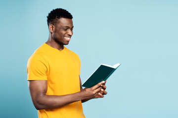 Portrait of handsome smiling African American man wearing yellow t shirt reading book isolated on blue background. Smart successful university student studying, learning language, education concept 