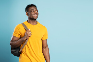 Handsome smiling African American student holding backpack looking away isolated on blue background, education concept. Portrait of happy handsome tourist