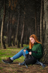 Hiker redhead woman pulling a canteen from backpack sitting under tree in forest. Trekking concept.