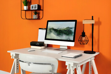 Modern computer, books, lamp and notebook on wooden desk near orange wall. Home office