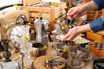 Woman holding vintage metal cup near table with different stuff, closeup. Garage sale