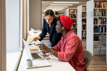 Concentrated diverse couple of students working on laptop together on study project in university library. Serious college teacher pointing out on computer screen mistake in solution to student guy