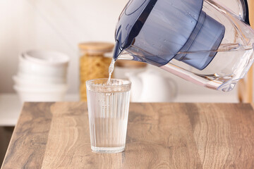 Pouring of pure water from filter jug into glass on table in kitchen