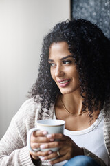 Looking out at the world. a cheerful young woman enjoying a cup of coffee while being seated on a chair at home during the day.