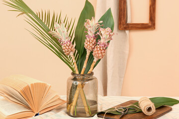Vase with decorative pineapples, palm leaves, book and rope on table near beige wall
