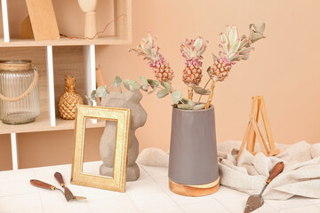 Vase with decorative pineapples, eucalyptus branches, frame and spatulas on table in room