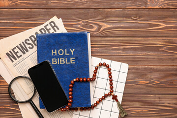 Holy Bible with prayer beads, mobile phone, magnifier, newspaper and notebook on wooden background