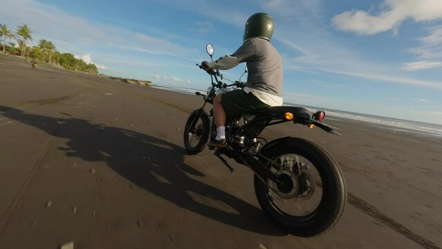 Handsome man biker driving his motorcycle cafe racer on the beach along the ocean during sunset. FPV footage