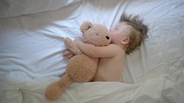 Funny baby sleeping on bed at home. Children's day lower sleeping position Sleeping baby happy and carefree in bed hugging a teddy bear toy. Mother cover with a blanket her child. Happiness in sleep