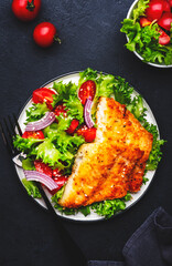 Fried white fish fillet with vegetable salad from lettuce, cherry tomatoes and red onion, black table background, top view