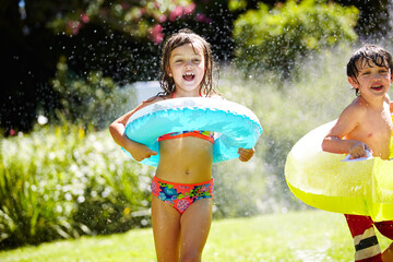 Cooling down and loving it. a brother and sister having fun with a sprinkler in the backyard. - obrazy, fototapety, plakaty