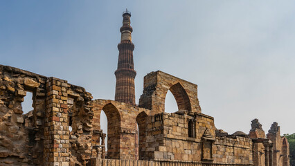 Ruins of the ancient temple complex Qutab Minar. Dilapidated walls with arches, carved decorations. Pigeons sit on ledges. The world's tallest brick minaret stands against the blue sky. India. Delhi