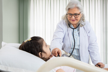 Obraz premium Indian senior female doctor pediatrician holding stethoscope checking heartbeat and breathing of sick girl child patient at hospital bed. healthcare concept. Closeup