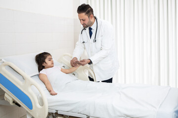 Obraz premium Young Indian male doctor pediatrician holding hand checking pulse of sick girl child patient at hospital bed. healthcare concept.