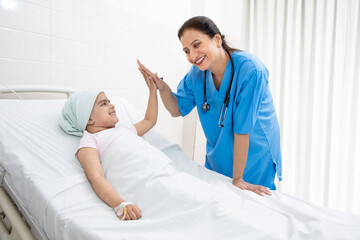 Happy indian woman nurse or medical staff give a high five to little girl cancer patient lying on...