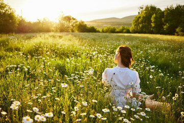 a red-haired woman in a light dress sits in a chamomile field at sunset and admires the passing day