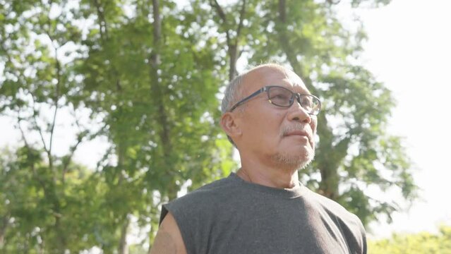 Active Asian senior man with eyeglasses is jogging at park outdoor. Healthy retirement lifestyle.