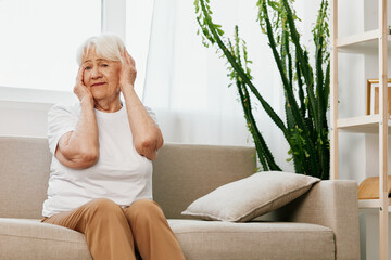 Elderly woman severe pain in the head sitting on the couch, health problems in old age, poor quality of life. Grandmother with gray hair holding her head, migraine and high blood pressure