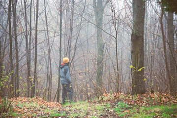 A young guy 30 y old with a yellow cap is walking in the forest. A subtle fog is in the wood. The guy is looking at something amongst the trees. 