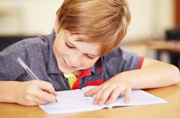 Naklejka premium School, education and a student boy writing in a book while sitting at his desk in a class room for learning. Kids, study and notebook with a young male pupil in a classroom for child development