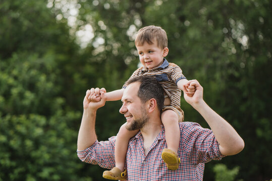 father rolls his little son on his shoulders. dad and child play and laugh in the garden on a warm summer day. a bearded man in a plaid shirt and a little boy roll around his neck