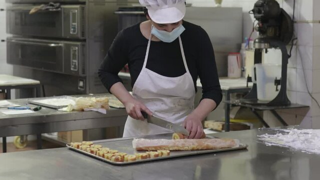Woman with mask cutting sweets on work table in bakery