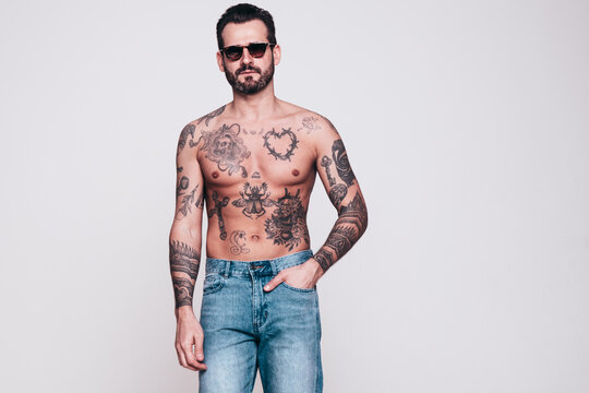 Portrait of handsome confident stylish hipster lambersexual model in jeans. Sexy modern man. Naked torso with tattoos. Fashion male posing in studio. Isolated on white. In sunglasses