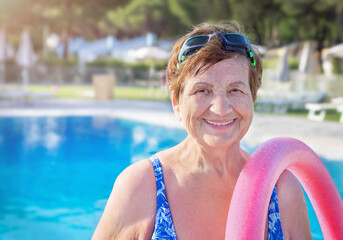 Active senior woman (over age of 50) in sport goggles, swimsuit and with swim noodles near swimming...