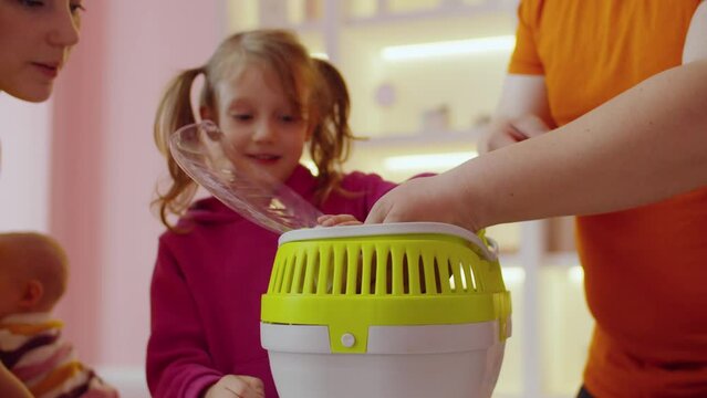 Close-up of curious fluffy white domestic rat looking out from pet carrier, while caring attractive parents introducing adopted pet to happy little girl ay home.