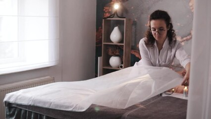 A cosmetologist or massage therapist prepares their workspace by covering the bed with a new disposable sheet for the next clients in a beauty salon with a stylish interior